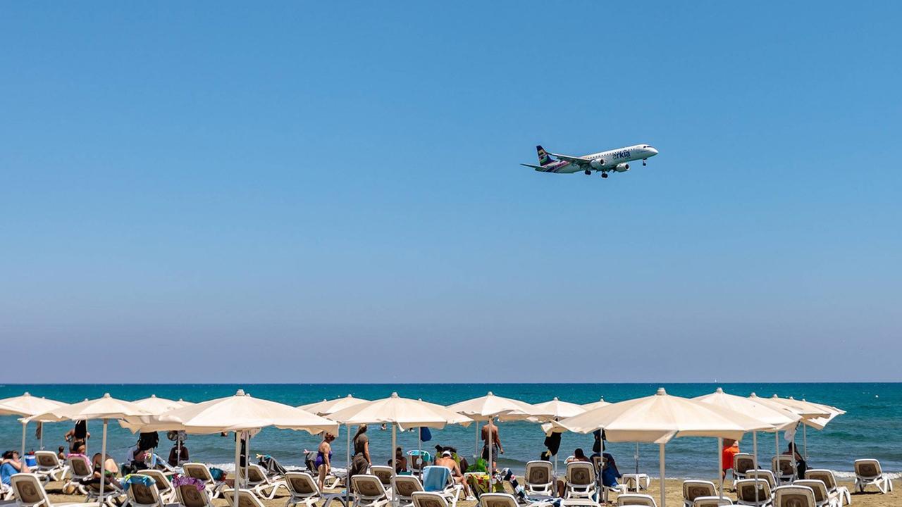 Ein Flugzeug fliegt bei strahlendem Sonnenschein tief über einem Strand mit Reihen voller Liegen und Touristen. Ein Flugzeug fliegt bei strahlendem Sonnenschein tief über einem Strand mit Reihen voller Liegen und Touristen.