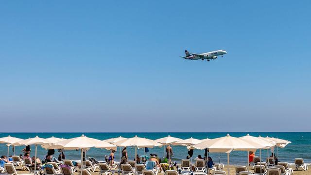 Ein Flugzeug fliegt bei strahlendem Sonnenschein tief über einem Strand mit Reihen voller Liegen und Touristen.