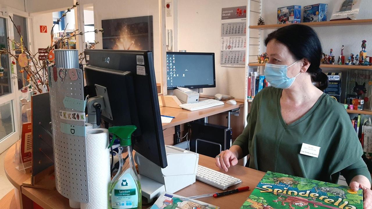 Eine Frau mit Schutzmaske steht in einer Bibliothek an einem Rechner. Eine Frau mit Schutzmaske steht in einer Bibliothek an einem Rechner.