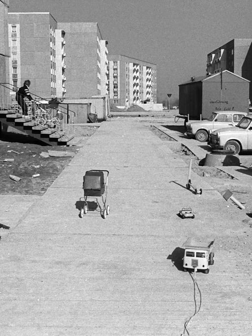Schwarzweiß Aufnahme eines Neubaugebietes — auf dem Weg vor den Häusern stehen ein Puppenwagen und ein kleiner Laster. Im Sand spielt ein Kind.