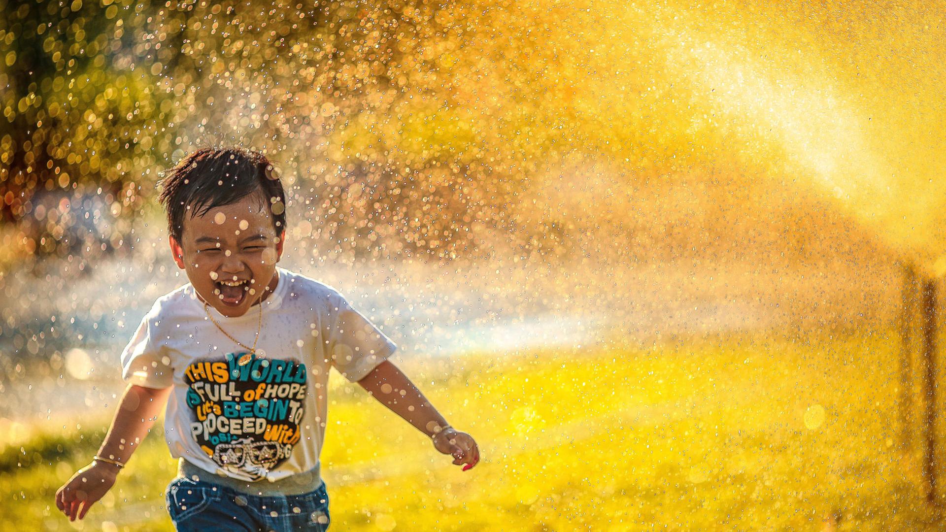 Ein kleiner Junge rennt begeistert unter dem Wassernebel einer Sprenkleranlage her Ein kleiner Junge rennt begeistert unter dem Wassernebel einer Sprenkleranlage her
