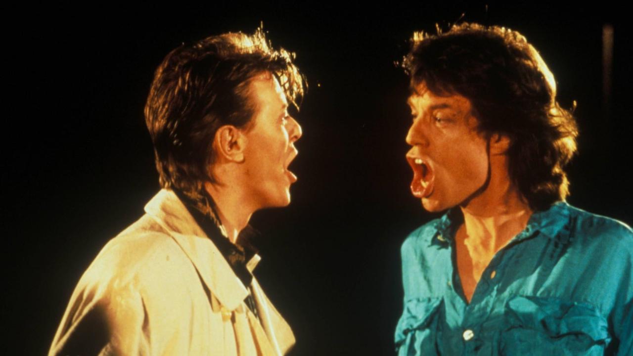 David Bowie links in heller Jacke und Mick Jagger rechts in türkisem Hemd sind im Profil zu sehen. Beide haben den Mund offen und schauen sich an, während sie "Dancing in the street" singen. David Bowie links in heller Jacke und Mick Jagger rechts in türkisem Hemd sind im Profil zu sehen. Beide haben den Mund offen und schauen sich an, während sie "Dancing in the street" singen.