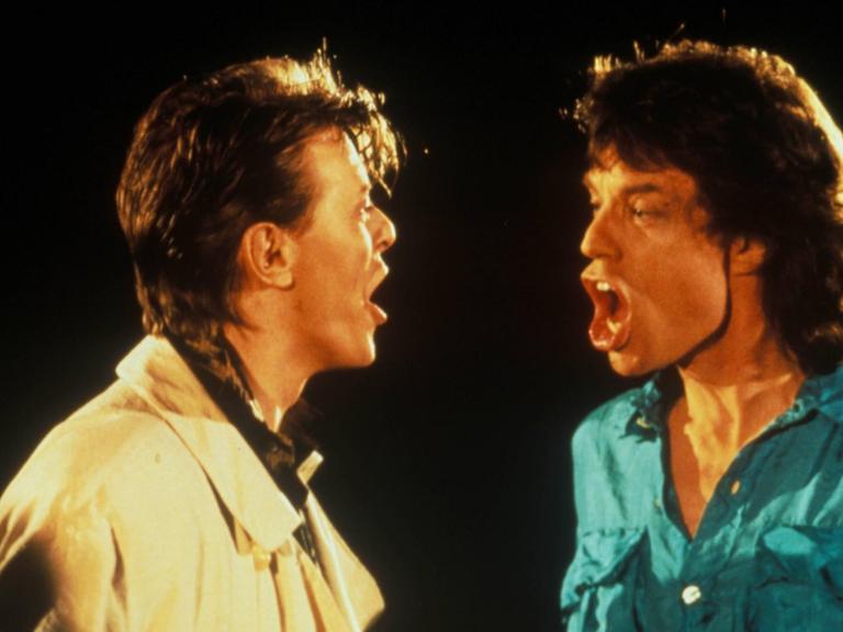 David Bowie links in heller Jacke und Mick Jagger rechts in türkisem Hemd sind im Profil zu sehen. Beide haben den Mund offen und schauen sich an, während sie "Dancing in the street" singen.