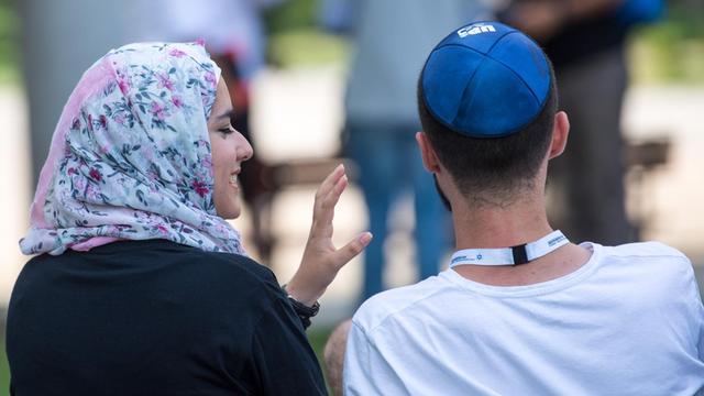 Eine junge Muslima mit Kopftuch und ein junger jüdischer Mann mit Kippa unterhalten sich miteinander.