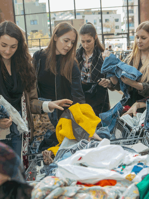 Auf einer Kleidertauschparty: Junge Frauen durchsuchen zwei Einkaufswagen nach Kleidungsstücken.