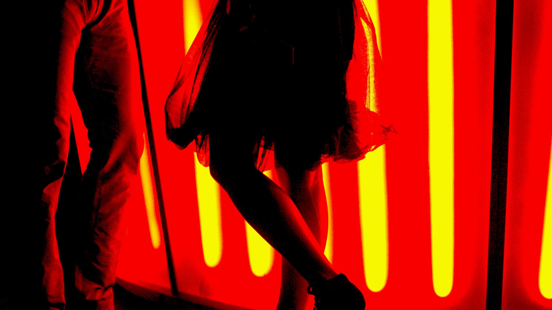Eine Frau mit einem teilweise transparenten Rock steht mit High-Heels an einer rot beleuchteten Bar und unterhält sich mit einem Mann. Eine Frau mit einem teilweise transparenten Rock steht mit High-Heels an einer rot beleuchteten Bar und unterhält sich mit einem Mann.
