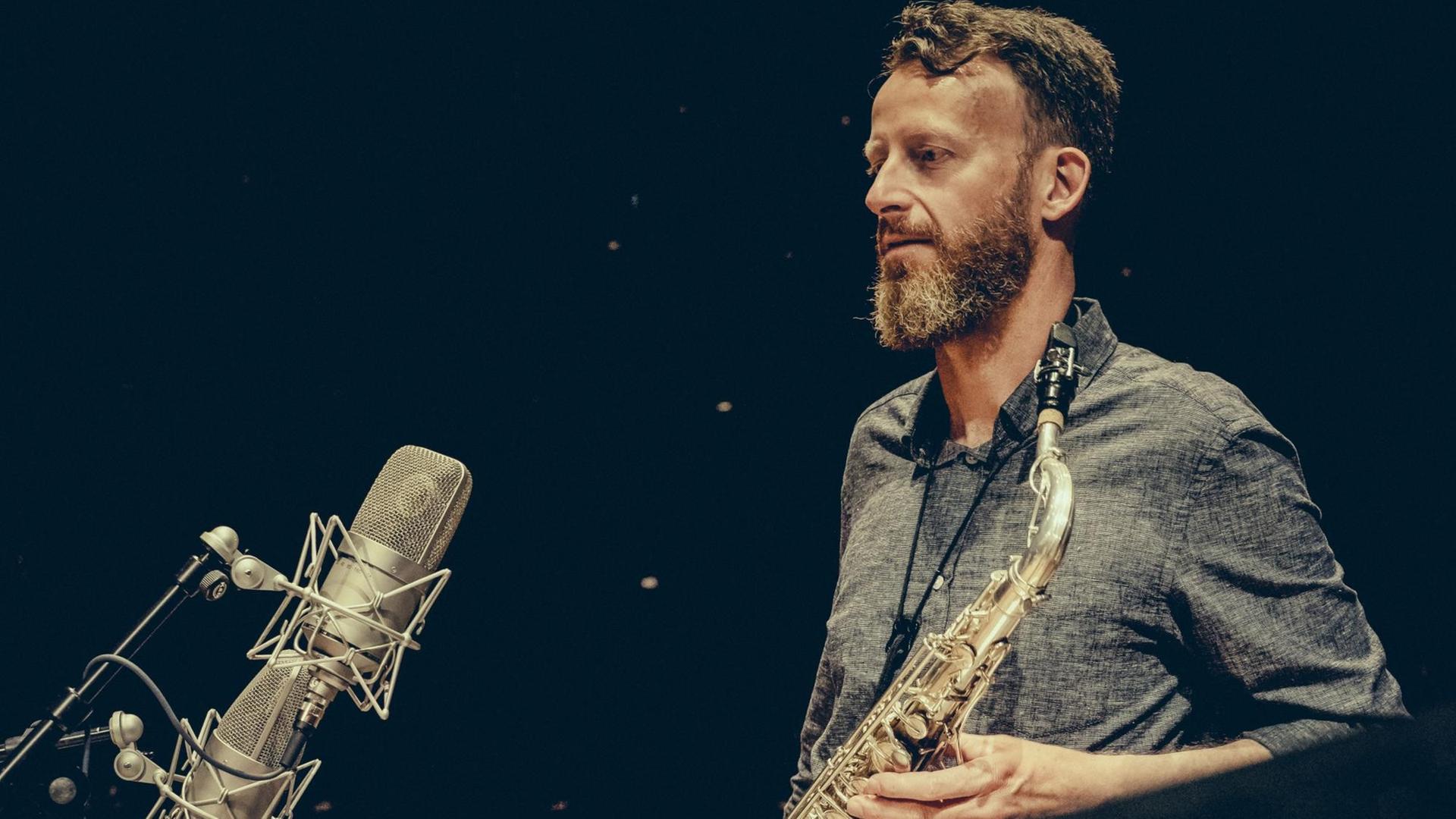 Ein Mann mit langen Bart und einem Saxofon in der Hand schaut mit verträumtem Blick auf die vor ihm aufgestellten Mikrofone.