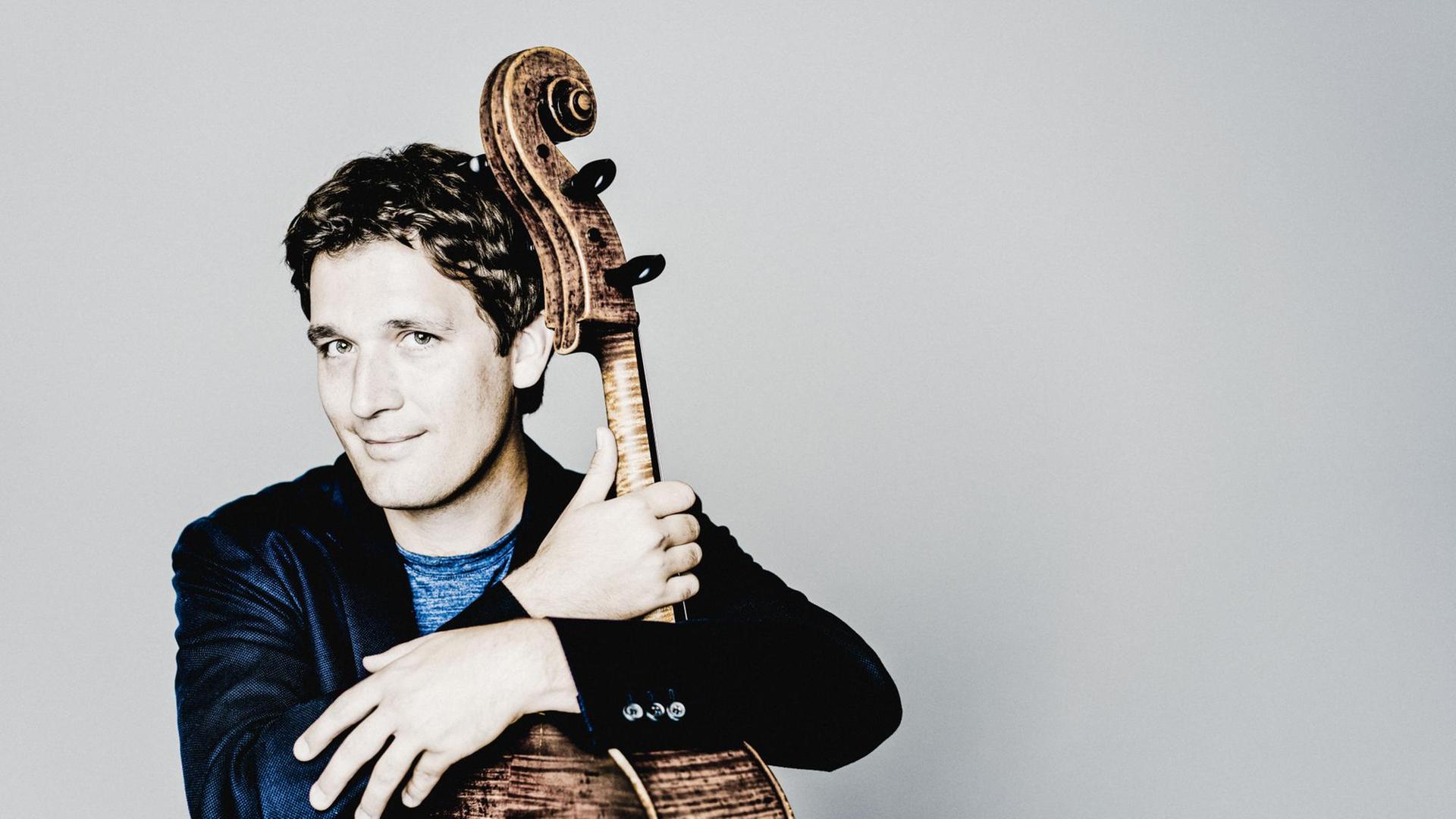 Maximilian Hornung hat die Arme um sein Cello gelegt und schaut lächelnd in die Kamera vor einem grauen Hintergrund. Maximilian Hornung hat die Arme um sein Cello gelegt und schaut lächelnd in die Kamera vor einem grauen Hintergrund.