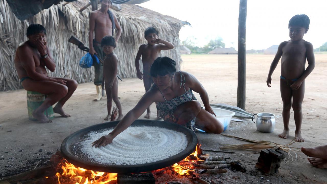 Eine Mutter bereitet im brasilianischen Dorf Kamayura im Xingu Nationalpark für ihre Kinder das aus Maniok hergestellte Lebensmittel Beiju zu. Dabei schauen ihr die Kinder und ein Mann mit einem Gewehr zu. Eine Mutter bereitet im brasilianischen Dorf Kamayura im Xingu Nationalpark für ihre Kinder das aus Maniok hergestellte Lebensmittel Beiju zu. Dabei schauen ihr die Kinder und ein Mann mit einem Gewehr zu.