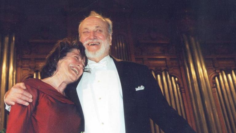 Ein Dirigent und eine Frau in rotem Kleid umarmen sich glücklich lachend auf einem Konzertpodium.