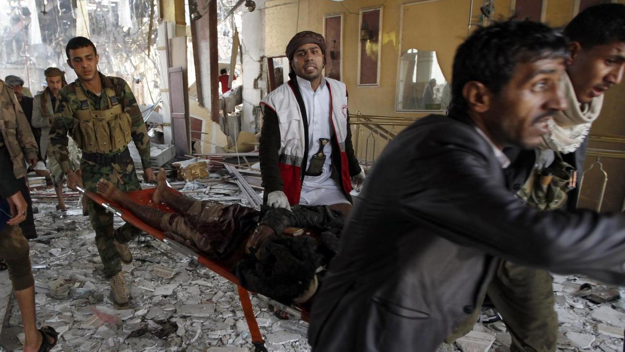 Drei Männer tragen das Opfer auf einer Bahre aus der zerstörten Halle, sie werden von einem Arzt begleitet. Andere Menschen schauen von der Seite zu. Der Boden ist mit Trümmern übersät.