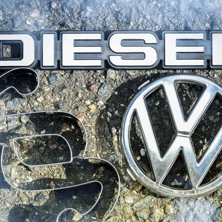 Ein VW-Logo, Paragrafen- und Dieselzeichen liegen auf der Straße. 