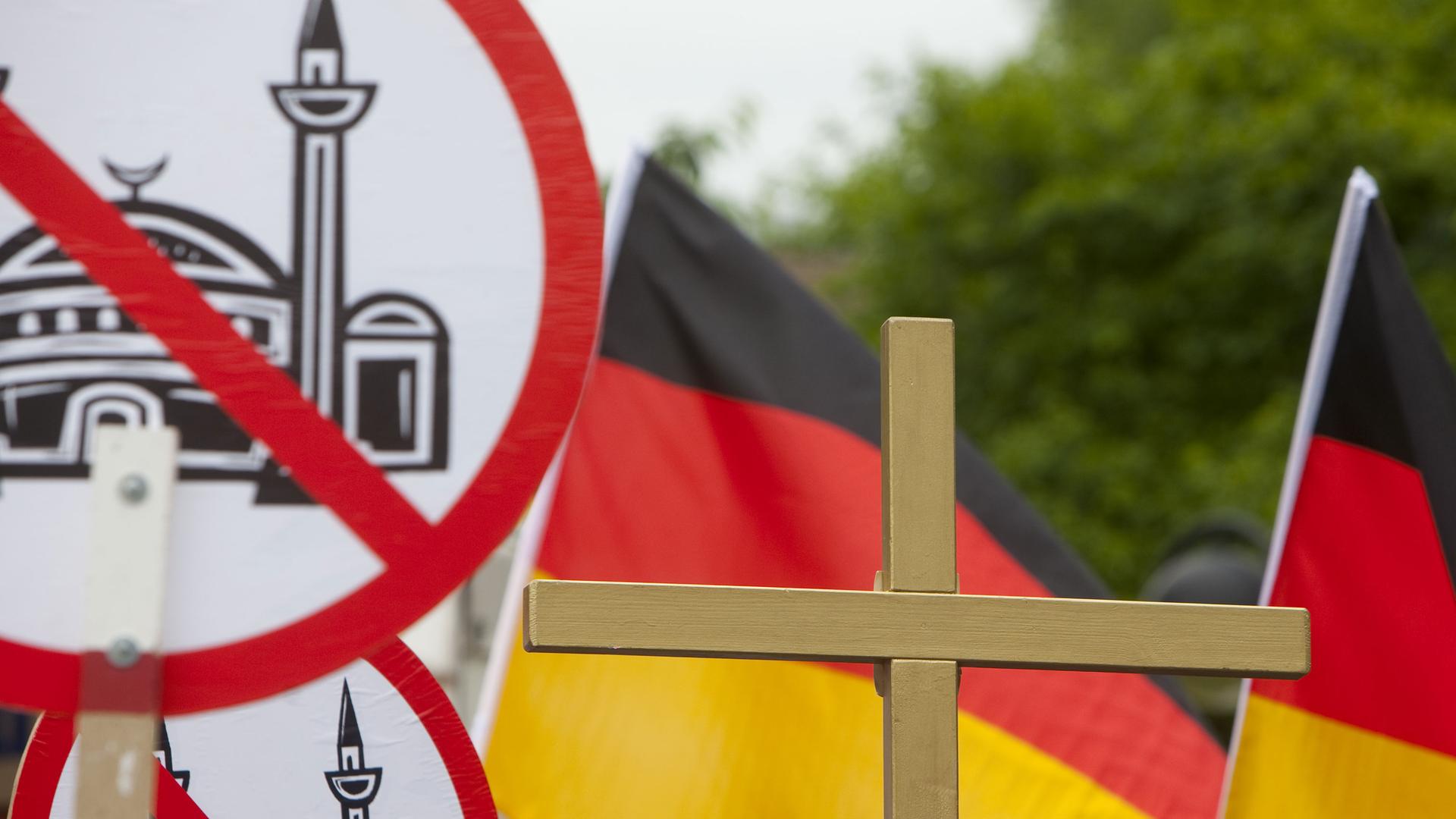 Auf dem Bild sind Schilder mit einer durchgestrichenen Moschee zu sehen, außerdem Deutschlandflaggen und ein Kreuz.