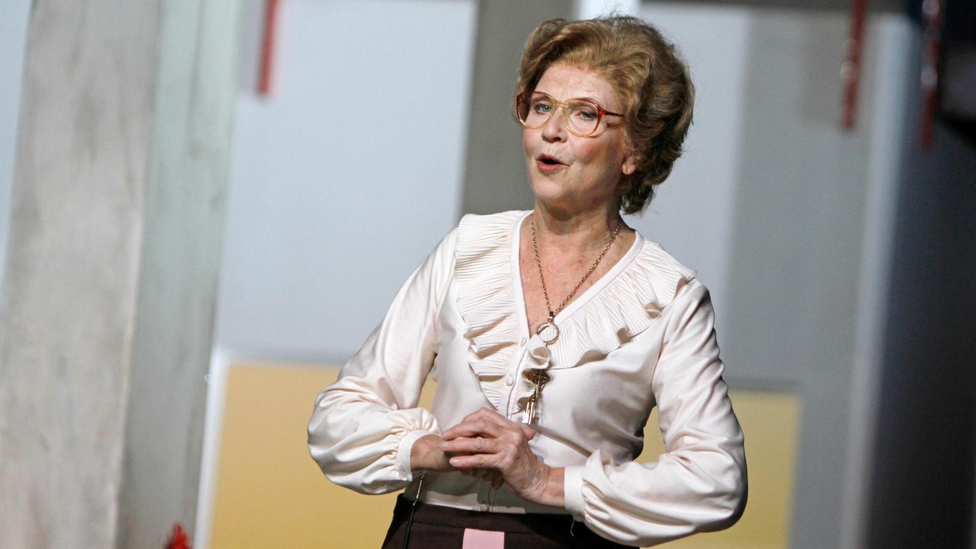 Zu sehen ist die Schauspielerin Irm Hermann, die auf einer Theaterbühne steht. Sie trägt ein Kleid und hält die Hände vor ihrem Körper.