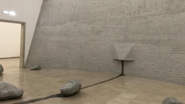 Installationsansicht: Steinartige Gebilde liegen vor einer grauen, betonartigen Wand, aus der eine nasenförmige Auswölbung ragt.