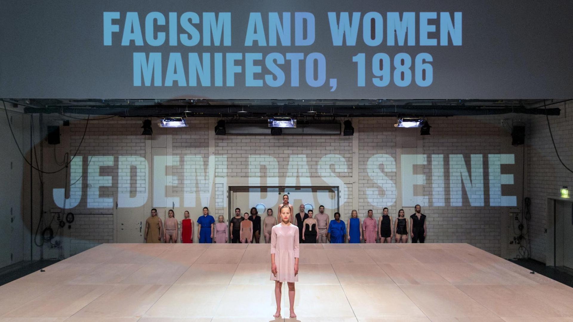 Eine Frau steht auf einer Theaterbühne, im Hintergrund stehen die Wörter: "Facism and Woman Manifesto, 1986" und "Jedem das seine" Eine Frau steht auf einer Theaterbühne, im Hintergrund stehen die Wörter: "Facism and Woman Manifesto, 1986" und "Jedem das seine"