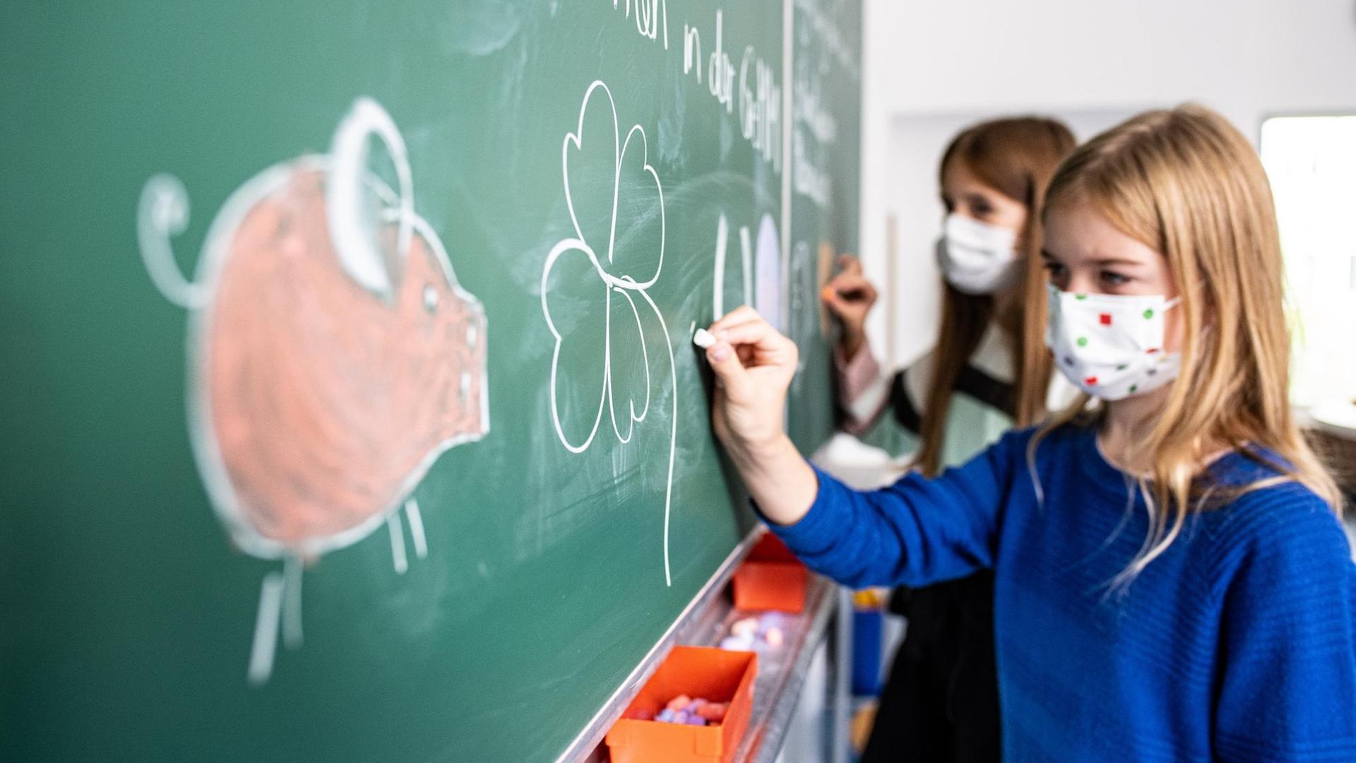 Zwei Schülerinnen mit Schutzmaske malen ein Schwein, Kleeblatt, Hufeisen und einen Luftballon an die Tafel in einem Klassenzimmer. Zwei Schülerinnen mit Schutzmaske malen ein Schwein, Kleeblatt, Hufeisen und einen Luftballon an die Tafel in einem Klassenzimmer.