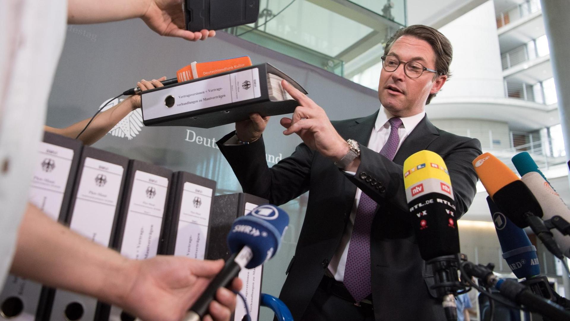 Scheuer hält einen Aktenordner hoch und zeigt mit dem Zeigerfinger darauf. Um ihn herum werden Mikrofone der Sender gehalten. Scheuer hält einen Aktenordner hoch und zeigt mit dem Zeigerfinger darauf. Um ihn herum werden Mikrofone der Sender gehalten.