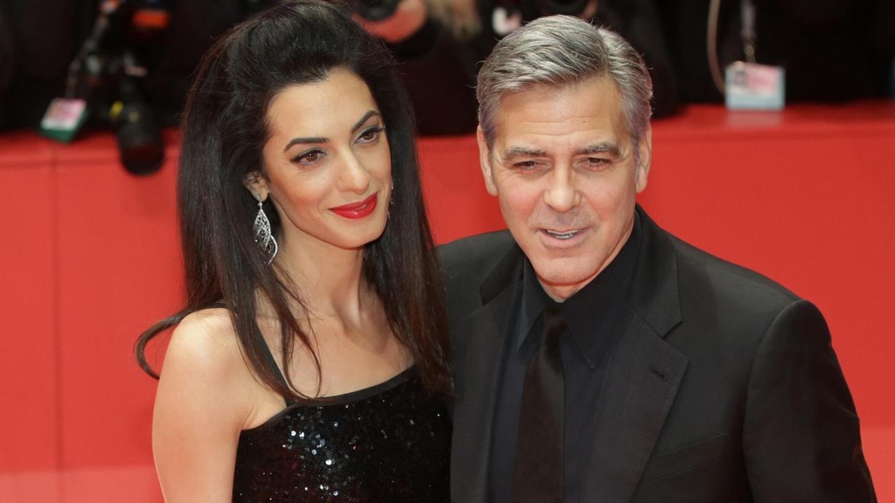 Clooney und Amal lächeln auf dem roten Teppich engumschlungen in die Kameras. Er trägt einen schwarzen Anzug, sie ein schwarzes schulterfreies Kleid.