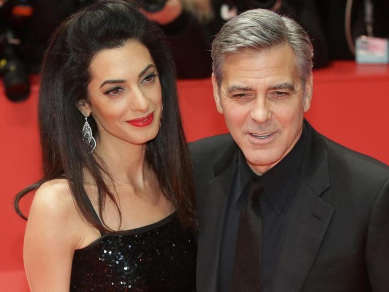Clooney und Amal lächeln auf dem roten Teppich engumschlungen in die Kameras. Er trägt einen schwarzen Anzug, sie ein schwarzes schulterfreies Kleid.