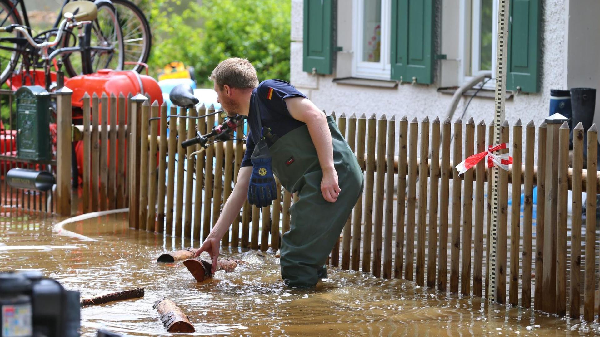 Ein Mitarbeiter des Technischen Hilfswerks steht knietief im schlammigen Wasser, hinter ihm ein Gartenzaum und ein Wohnhaus. Gegenstände treiben durch das Wasser. Ein Mitarbeiter des Technischen Hilfswerks steht knietief im schlammigen Wasser, hinter ihm ein Gartenzaum und ein Wohnhaus. Gegenstände treiben durch das Wasser.