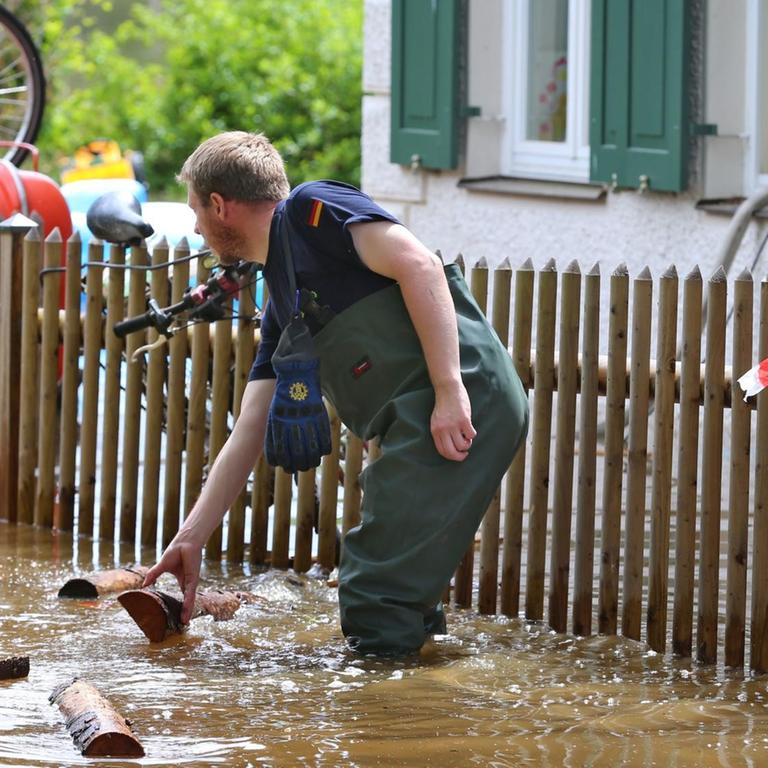 Ein Mitarbeiter des Technischen Hilfswerks steht knietief im schlammigen Wasser, hinter ihm ein Gartenzaum und ein Wohnhaus. Gegenstände treiben durch das Wasser.