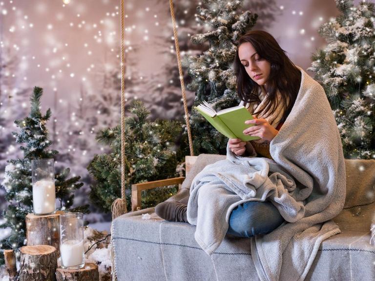 Eine junge Frau sitzt vor romantischer Weihnachtskulisse und liest ein Buch.