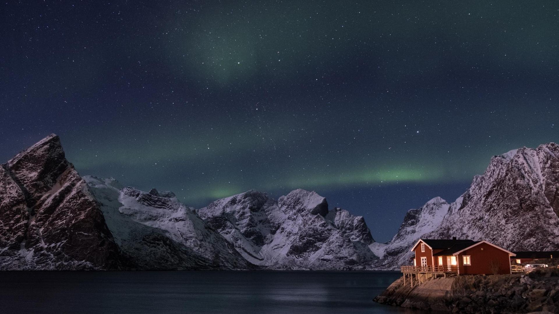 Nächtliche Szene eines erleuchteten Holzhäuschens an der norwegischen Küste mit Bergen und Polarlichtern im Hintergrund. Nächtliche Szene eines erleuchteten Holzhäuschens an der norwegischen Küste mit Bergen und Polarlichtern im Hintergrund.
