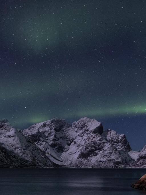 Nächtliche Szene eines erleuchteten Holzhäuschens an der norwegischen Küste mit Bergen und Polarlichtern im Hintergrund.