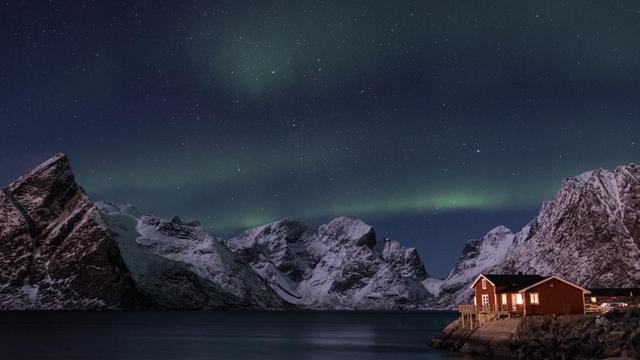 Nächtliche Szene eines erleuchteten Holzhäuschens an der norwegischen Küste mit Bergen und Polarlichtern im Hintergrund.