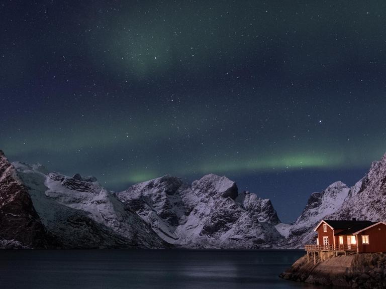 Nächtliche Szene eines erleuchteten Holzhäuschens an der norwegischen Küste mit Bergen und Polarlichtern im Hintergrund.