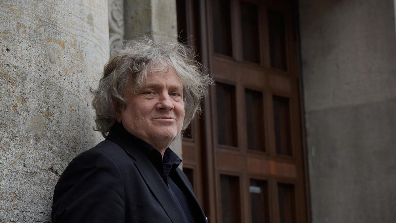 Carl Hegemann steht vor den Mauern der Volksbühne am Rosa-Luxemburg-Platz in Berlin. Er hat lockige, graue Haare und trägt dunkle Kleidung. Im Hintergrund ist das Eingangsportal des Theaterbaus zu sehen. Carl Hegemann steht vor den Mauern der Volksbühne am Rosa-Luxemburg-Platz in Berlin. Er hat lockige, graue Haare und trägt dunkle Kleidung. Im Hintergrund ist das Eingangsportal des Theaterbaus zu sehen.