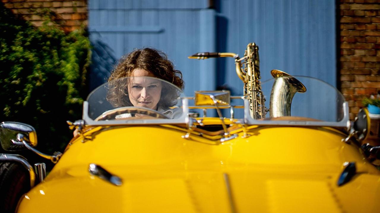 Die Musikerin sitzt in einem gelben Oldtimer, neben ihr ihr Instrument auf dem Beifahrersitz.