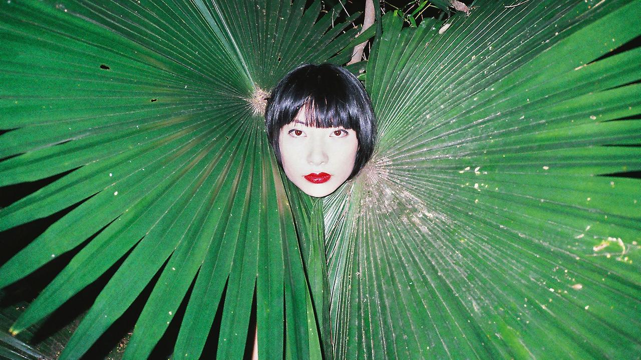 Eine Frau schaut in Mitten zweier Palmenblätter den Fotografen an, "Untitled, 2015" von Ren Hang. Eine Frau schaut in Mitten zweier Palmenblätter den Fotografen an, "Untitled, 2015" von Ren Hang.