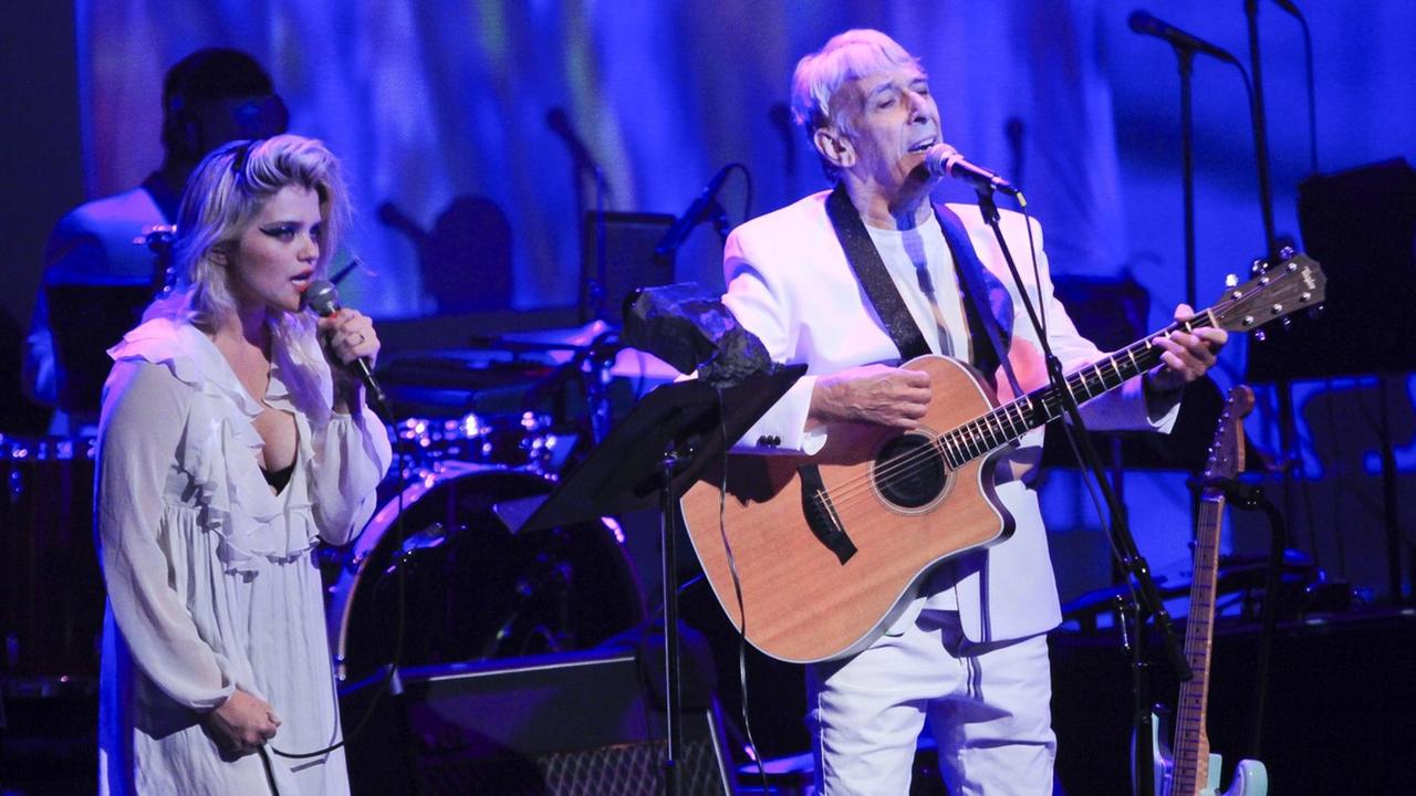 John Cale mit Gitarre auf einer in blaues Licht getauchten Bühne. Neben ihm steht die Sängerin Sky Ferreira. John Cale mit Gitarre auf einer in blaues Licht getauchten Bühne. Neben ihm steht die Sängerin Sky Ferreira.
