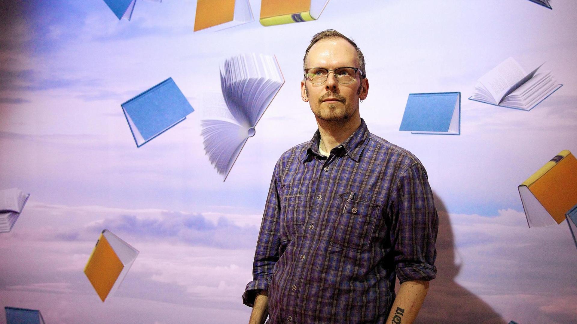 Dietmar Dath steht vor einer Wand. Auf dieser sind Bücher in verschiedenen Farben zu sehen, die durch die Luft segeln. Dietmar Dath steht vor einer Wand. Auf dieser sind Bücher in verschiedenen Farben zu sehen, die durch die Luft segeln.