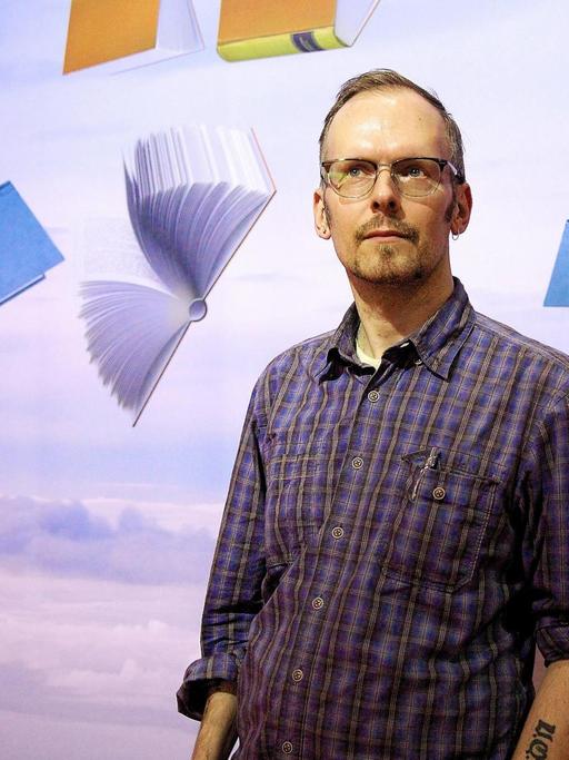 Dietmar Dath steht vor einer Wand. Auf dieser sind Bücher in verschiedenen Farben zu sehen, die durch die Luft segeln.