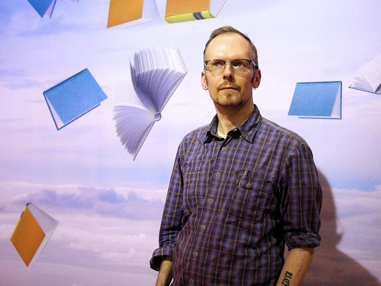 Dietmar Dath steht vor einer Wand. Auf dieser sind Bücher in verschiedenen Farben zu sehen, die durch die Luft segeln.