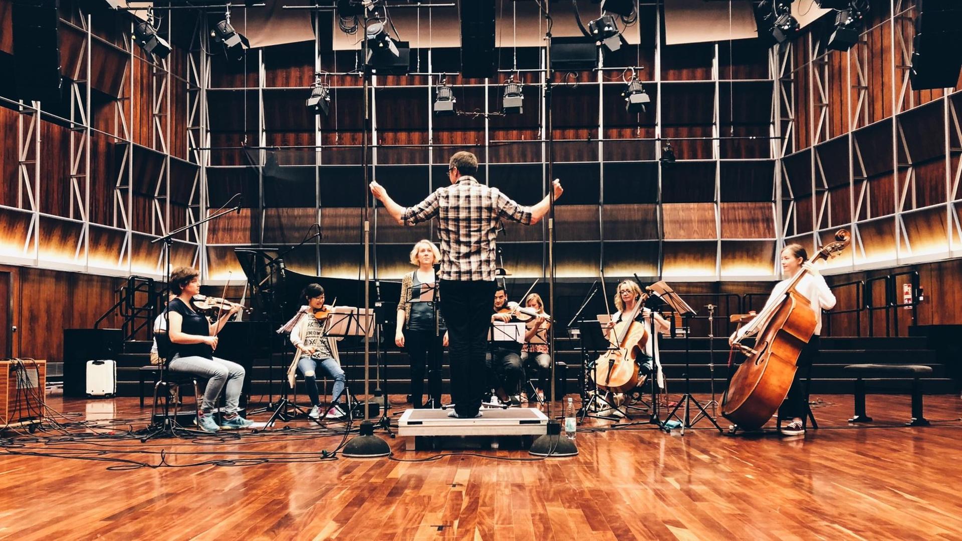Ein Dirigent steht auf einem Podest vor einer Gruppe Musikerinnen und Musiker, die Streichinstrumente spielen. Ein Dirigent steht auf einem Podest vor einer Gruppe Musikerinnen und Musiker, die Streichinstrumente spielen.