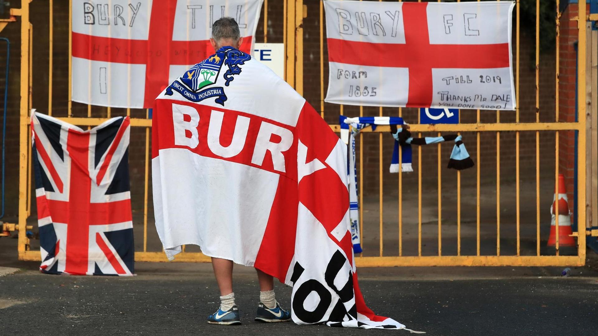 Ein Fan mit FC-Bury-Flagge um die Schultern gebunden steht vor dem Zaun des Stadions. Ein Fan mit FC-Bury-Flagge um die Schultern gebunden steht vor dem Zaun des Stadions.