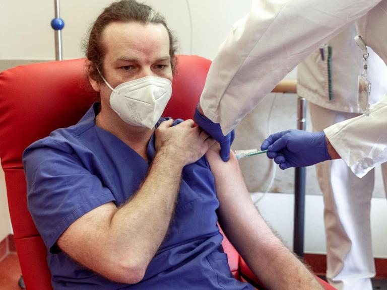 Ein Pfleger in blauem Krankenhauskittel sitzt auf einem roten Stuhl und bekommt von einem Arzt in weiß eine Impfung in den Oberarm gesetzt.