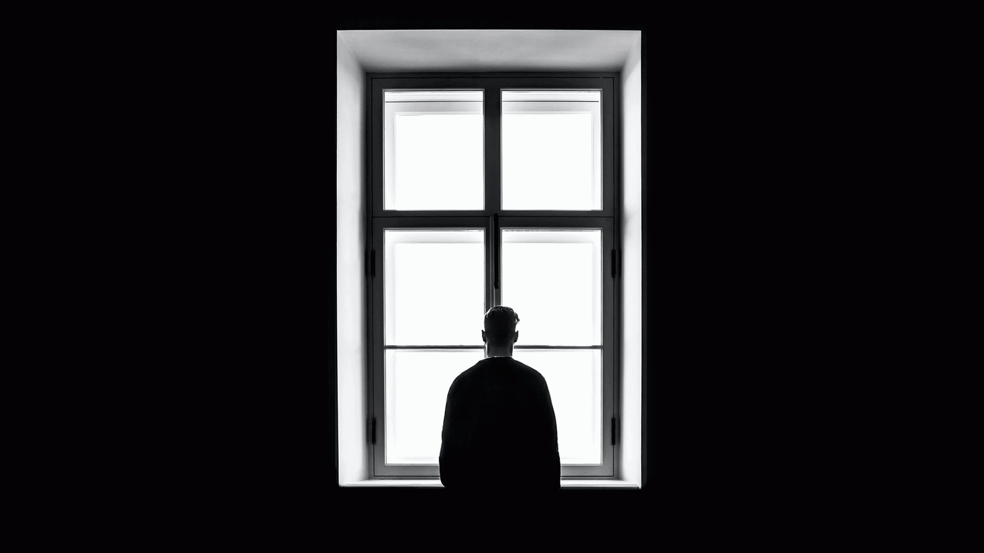 Ein Mann steht mit dem Rücken zur Kamera und schaut aus einem Fenster. Die Wände sind schwarz. Ein Mann steht mit dem Rücken zur Kamera und schaut aus einem Fenster. Die Wände sind schwarz.