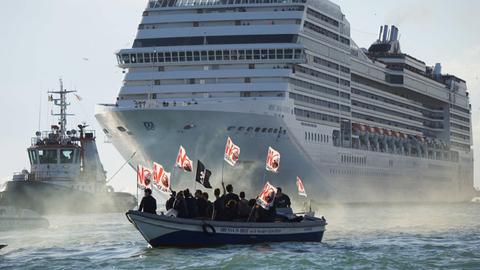 Als Silhouetten heben sich Demonstranten mit Plakaten in einem kleinen Boot vor einem riesigen Kreuzfahrtschiff ab.