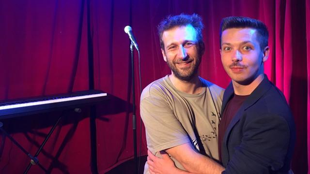 Mike Topolski (r.) aus Polen und Radu Isac aus Rumänien umarmen sich und stehen auf einer Bühne mit Mikrofon im Hintergrund