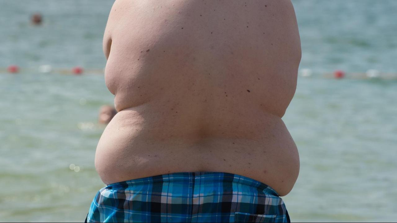 Ein übergewichtiger Junge in Badehose steht an einem Strand.