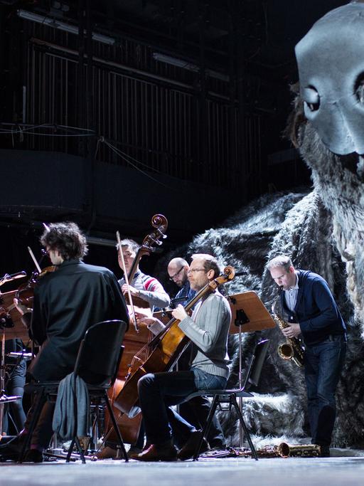 Eine überdimensionale Figur eines Hundes liegt in Bochum im Schauspielhaus während einer Probe auf der Bühne, während Musiker daneben sitzen.