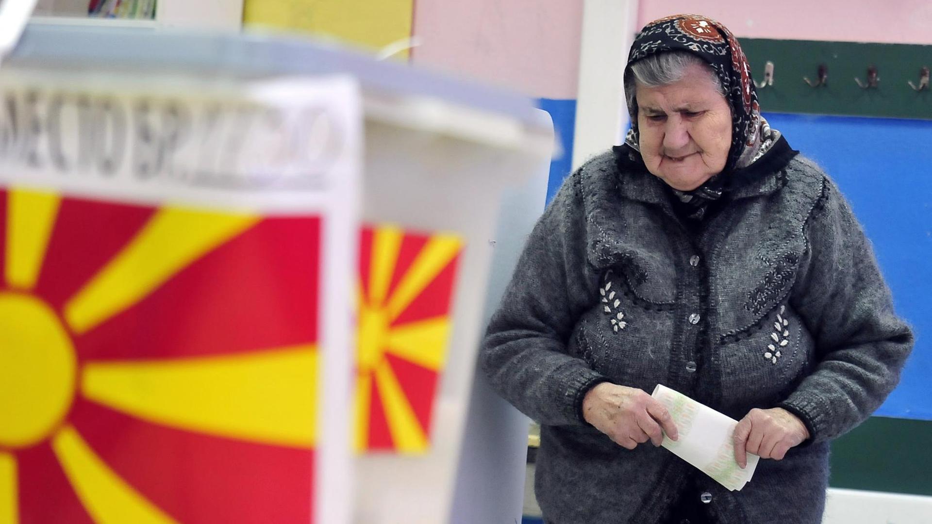Eine ältere Frau mit Kopftuch kommt aus einer Wahlkabine heraus, den Stimmzettel in der Hand.