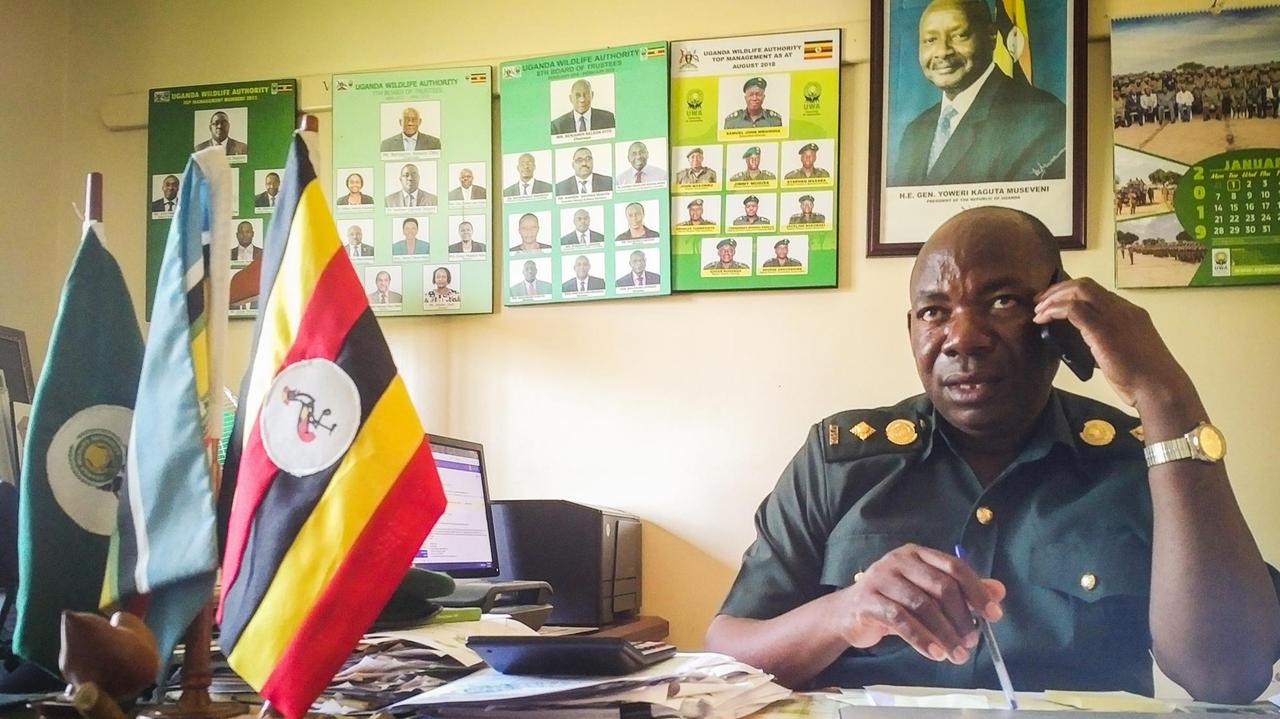 Der Chef-Ranger des Queen-Elizabeth-Nationalparks Edward Asalu sitzt in seinem Büro am Schreibtisch zwischen Fähnchen und Fotos höherer Beamter und des Präsidenten.