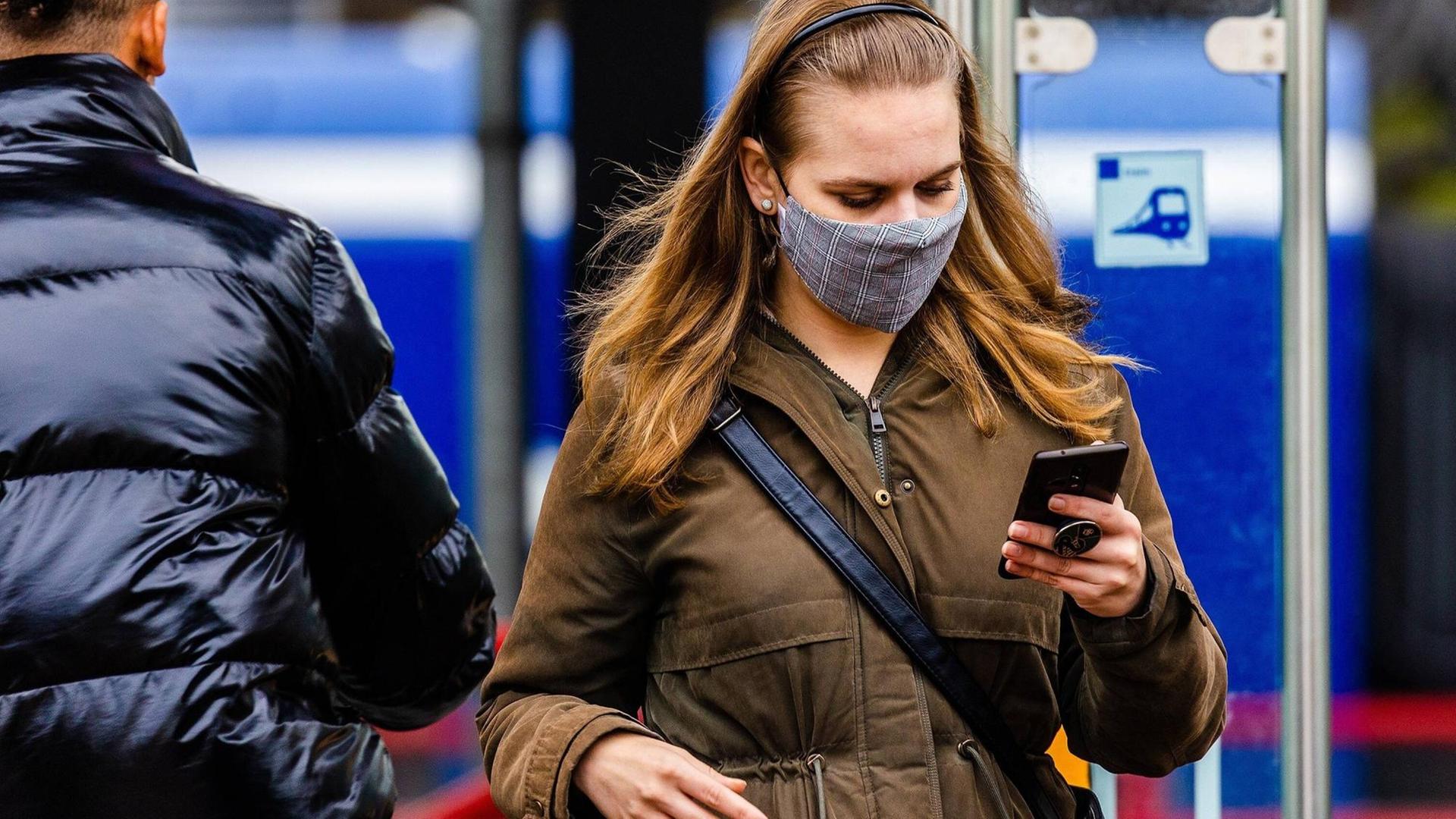 Junge Frau mit Maske in Dordrecht, Niederlande, schaut auf ihr Smartphone Junge Frau mit Maske in Dordrecht, Niederlande, schaut auf ihr Smartphone