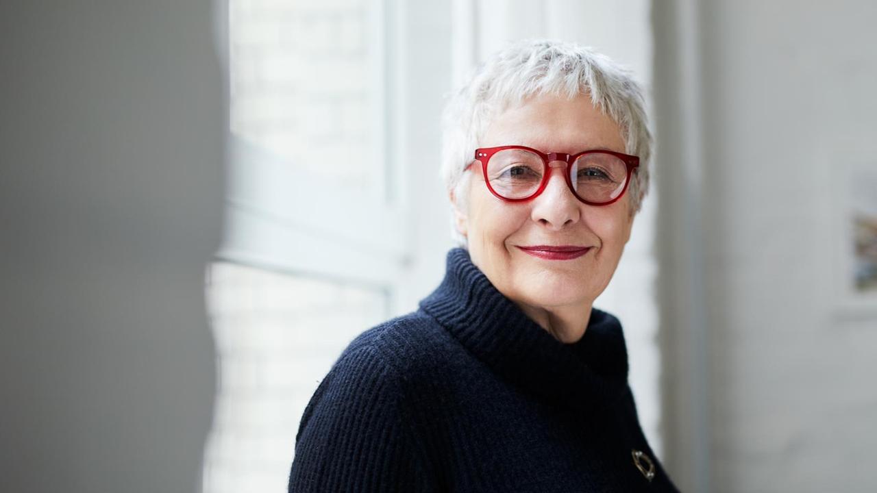 Erica Fischer blickt in einem Zimmer in die Kamera. Sie trägt ein dunkles Oberteil, hat grau-weiße Haare und trägt eine rote Brille. Erica Fischer blickt in einem Zimmer in die Kamera. Sie trägt ein dunkles Oberteil, hat grau-weiße Haare und trägt eine rote Brille.